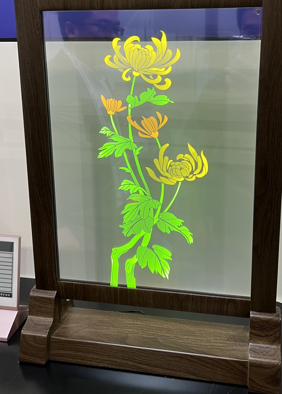 Transparent OLED panel in a wooden display stand, showing an emissive chrysanthemum with the background visible through the glass